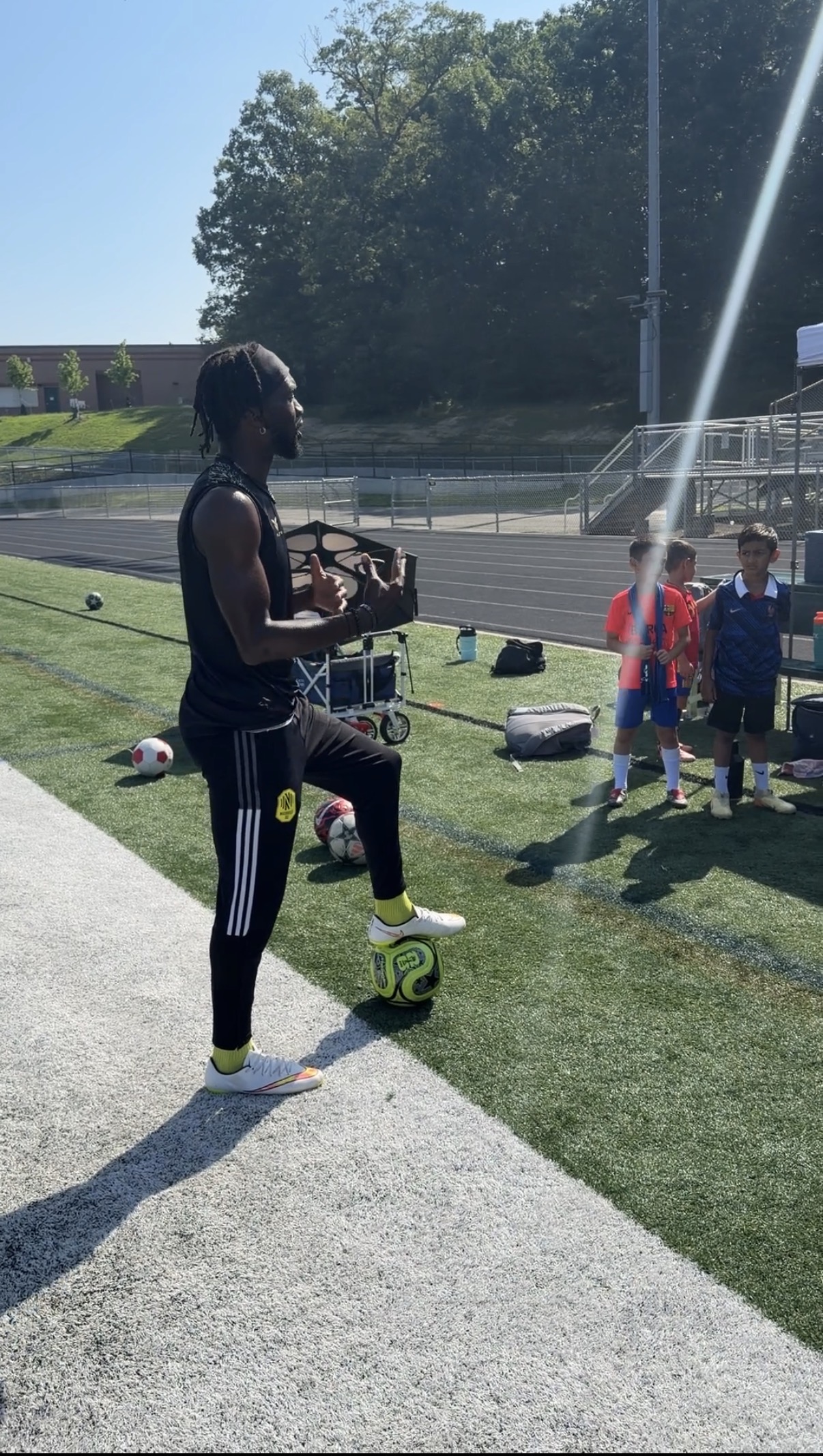 CJ Sapong holding a rebound net during a training session, young players waiting on the track behind him.